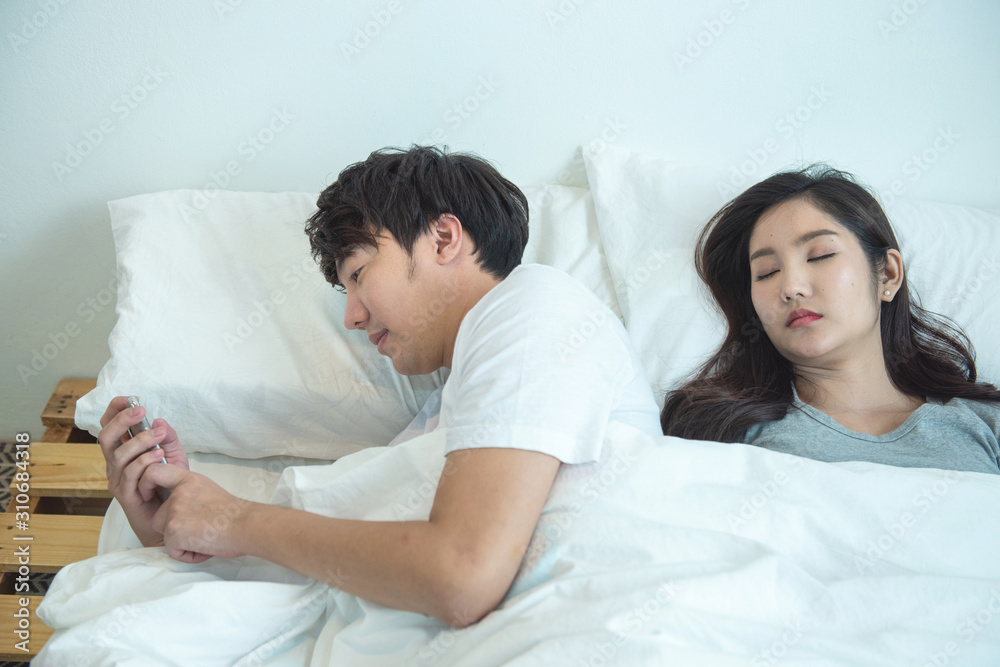 Fototapeta premium Asian young man use smart mobile phone chatting secrecy on bed while his girlfriend sleeping.Cheating boyfriend Secretly use cellphone,lifestyle relationship social problem.
