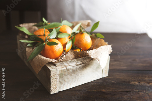 Fresh mandarin oranges fruits or tangerines with leaves in the wooden box on dark rustic table near window. Natural light, selective focus, copy space. 