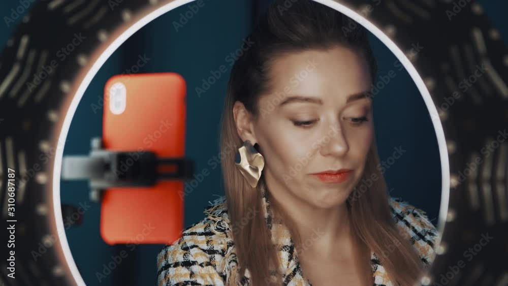 Confident pretty female video blogger sits in front of ring light and orange smartphone, applying red lipstick, recording tutorial video for beauty channel. Shot on 4K RED camera. Confident pretty female video blogger sits in front of ring light and orange smartphone, applying red lipstick, recording tutorial video for beauty channel. Shot on 4K RED camera.