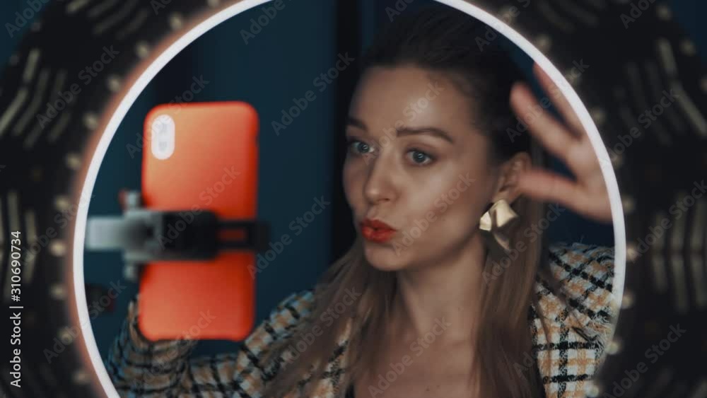 View of cute young blonde woman beauty blogger dancing in front of ring light, looking at camera of smartphone, recording tutorial video for channel. Shot on 4K RED camera. View of cute young blonde woman beauty blogger dancing in front of ring light, looking at camera of smartphone, recording tutorial video for channel. Shot on 4K RED camera.