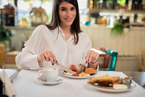 Beautiful caucasian elegant brunette sitting in restaurant and eating sausage for breakfast.