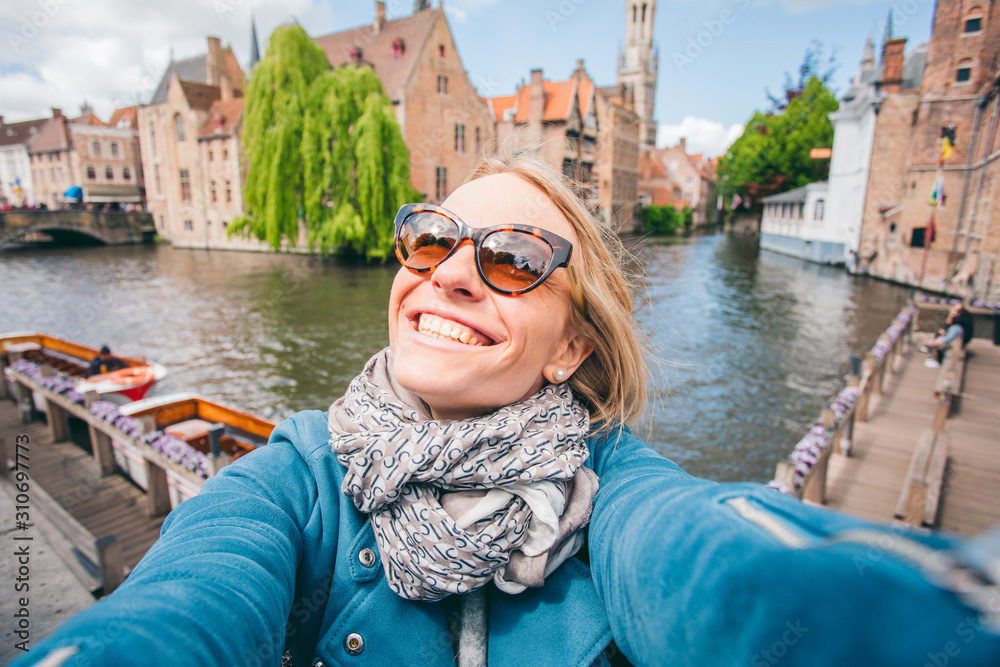 Beautiful young girl takes selfie photo on the background of the famous tourist destination with a canal in Bruges, Belgium