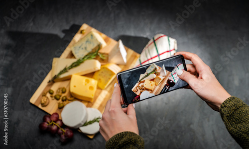 Hands taking photo a cheese platter with different cheese on a dark background with smartphone