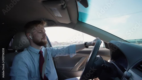 Side view shot of a stylish business man driving a car in city in summer. hand on steering wheel. listening to music or podcast in headphones airpods. driver, work, millenial, hipster concept