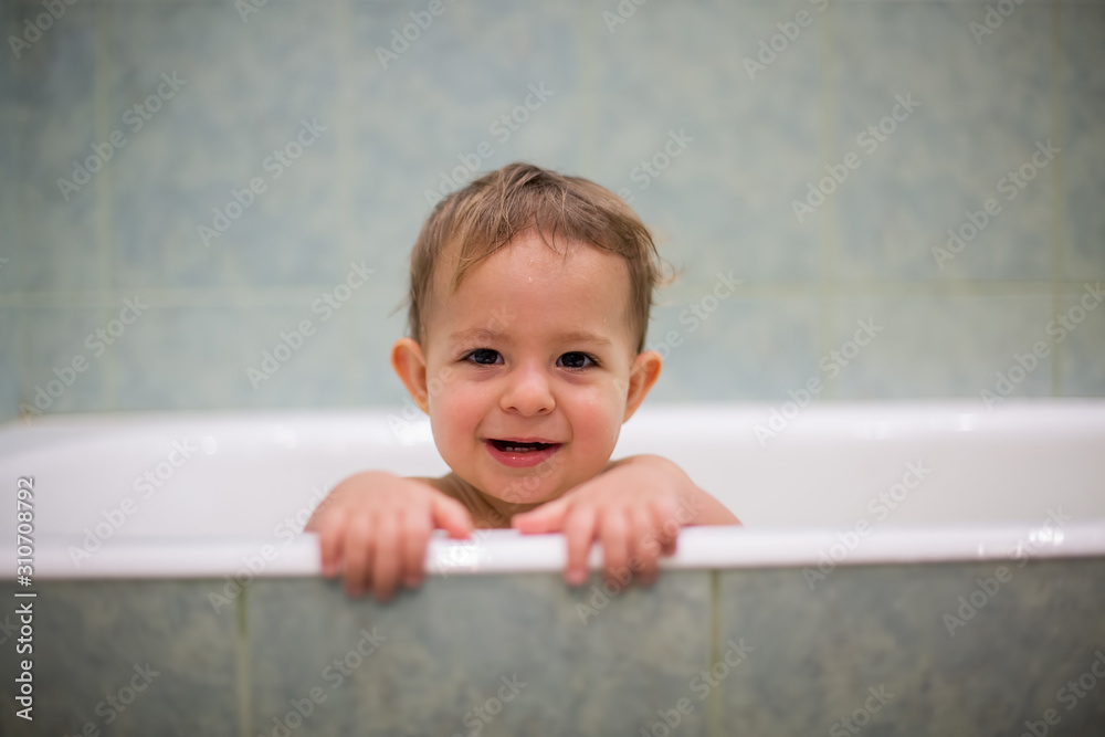 A cute Caucasian baby peeks out of the bathtub, put hands on the side of the bath and looks at the camera with smile and laugh. in the background is a green bathroom in blur. close-up, soft focus A cute Caucasian baby peeks out of the bathtub, put hands on the side of the bath and looks at the camera with smile and laugh. in the background is a green bathroom in blur. close-up, soft focus