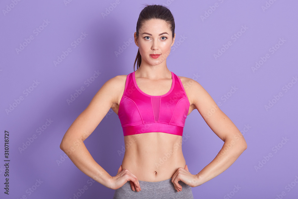 Winsome Caucasian woman with pink workout clothing and ponytail, posing isolated over lilac background, serious sporty female kepping hands on hips and looking directly at camera. Fitness concept.