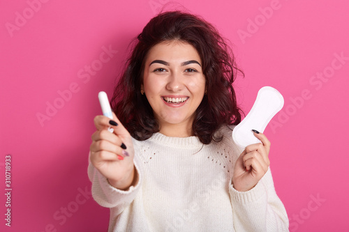 Happy satisfied Caucasian woman holding different types of feminine hygiene products during menstrual cycle  sanitary pad and cotton tampon in fwemales hands  standing isolated over pink background.