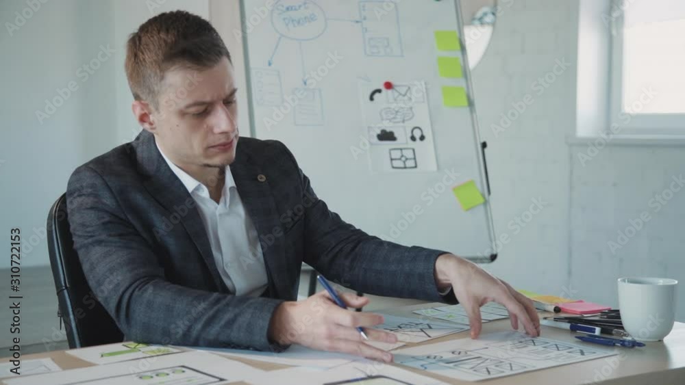 Qualified and professional graphic or web designer in formal wear clothes sitting behind table in coworking space with modern interior office. Man creating new website page and checking sketch