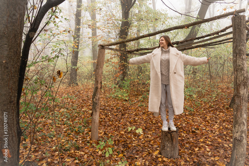 Happy smiling brunette girl in white coat stands with wide hands on the stump among the forest in fog photo