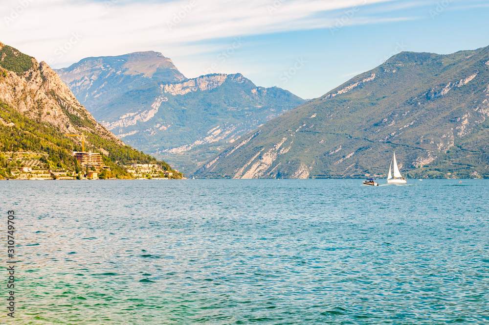 Beautiful Garda lake in Italy surrounded by high dolomite mountains. Classic white sailing yacht floating on the lake. Various hotels and private houses built on the rocky shores of the lake