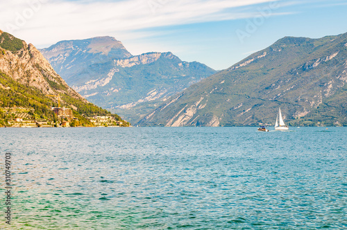 Beautiful Garda lake in Italy surrounded by high dolomite mountains. Classic white sailing yacht floating on the lake. Various hotels and private houses built on the rocky shores of the lake