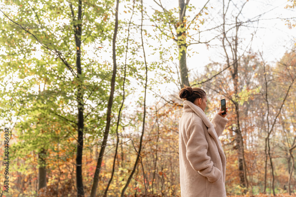 Attractive beautiful brunette girl in white coat walking among the colorful forest and making photo on her mobile phone Attractive beautiful brunette girl in white coat walking among the colorful forest and making photo on her mobile phone