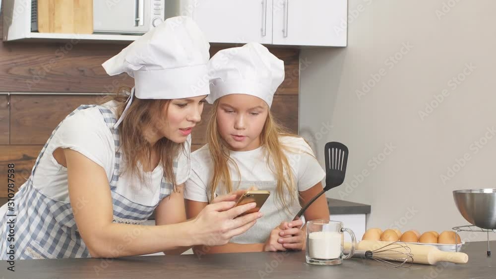 Happy family in aprons look at screen of smartphone watching delicious cake recipe, going to cook together. Happy family in aprons look at screen of smartphone watching delicious cake recipe, going to cook together.