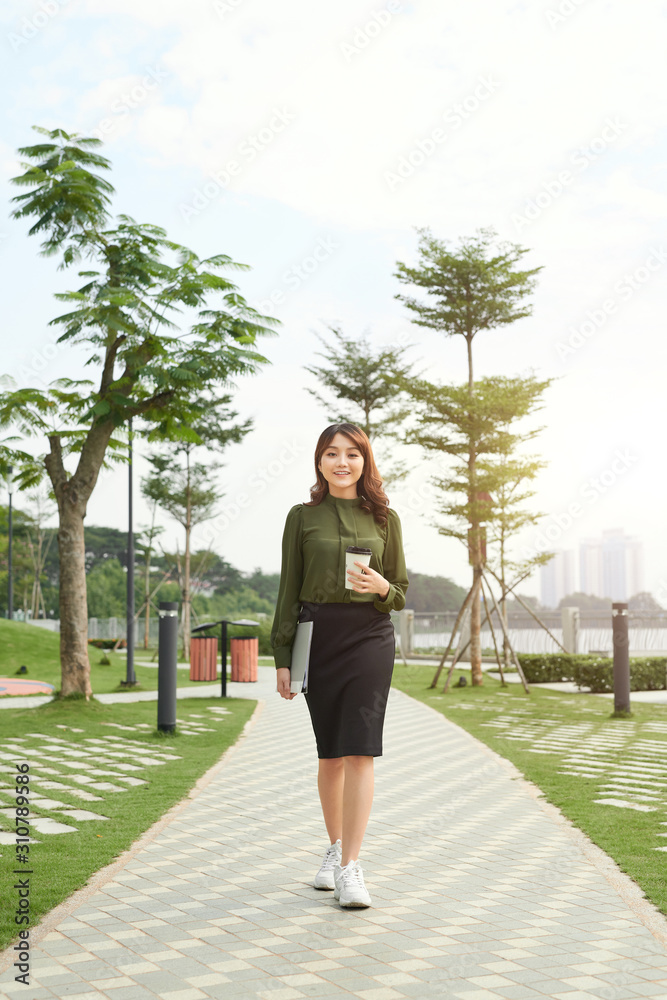 pretty young woman carrying a cup of takeaway coffee and a file in an urban park as she gives the camera a dazzling smile pretty young woman carrying a cup of takeaway coffee and a file in an urban park as she gives the camera a dazzling smile