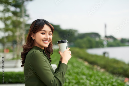 Nature, people concept - young asian woman standing in the park with a cup of coffee