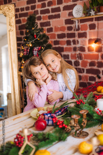 Cute sisters in pajamas have fun in the kitchen by the table with gingerbread cookies, tangerines at home in the room decorated with Christmas garlands, needles. Christmas mood