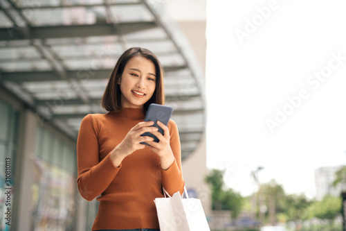 Fotografie Portrait of elegant young Asian woman holding shopping bags and using smartphone