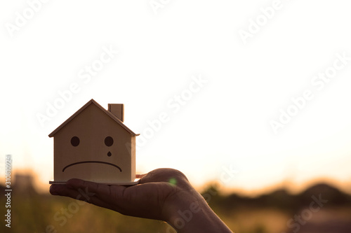 Closeup hand holding mockup house wood with face sad emotion at sunset background. Minimal design. House construction and decorate concept.