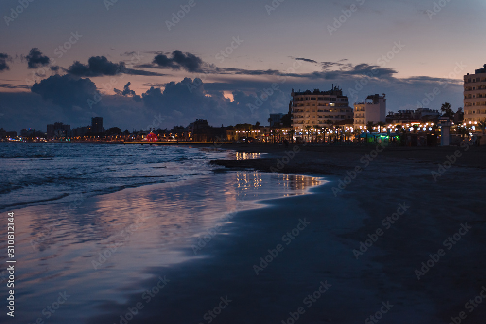 Evening beach at Cape Greco. Behind the dark silhouettes of people and houses lit by lights. Cyprus