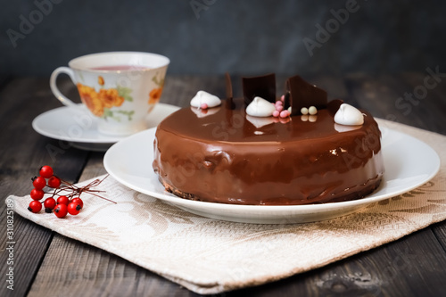 On a dark tabletop, on a linen napkin on a white dish, there is a round chocolate cake, next to a white cup is dark tea and next to the round berries