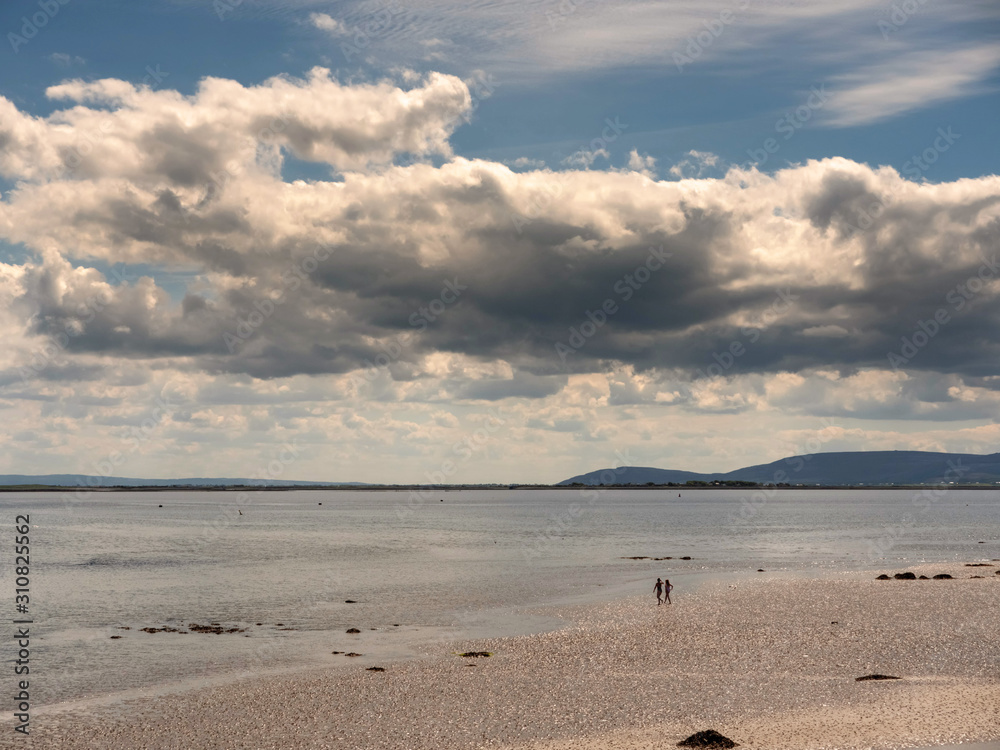 Silhouette of two woman on a sandy beach walking towards water, Galway bay, Burren mountains in the background, Cloudy sky over ocean.