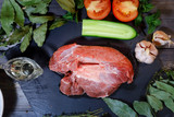 A piece of raw beef is laid out on a dark wooden tabletop on a graphite stone tile, next to a sauceboat with butter, peas, peppermint, laurel leaves, thyme, olives, garlic
