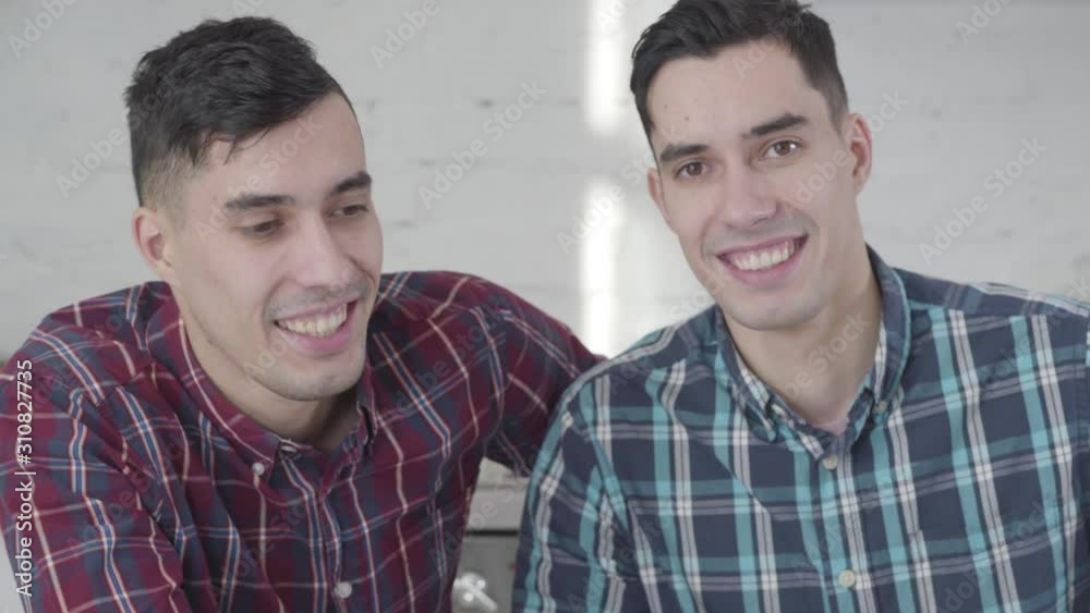 Close-up portrait of Caucasian twin brothers looking at camera and smiling. Two positive young men posing at home. Friendship, family, identical siblings. Close-up portrait of Caucasian twin brothers looking at camera and smiling. Two positive young men posing at home. Friendship, family, identical siblings.