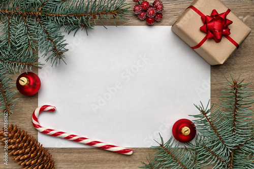 Christmas or New Year composition. White sheet of paper with copy space, gift box, candy cane, Christmas ball and fir tree branches on wooden background. Flat lay, view from above
