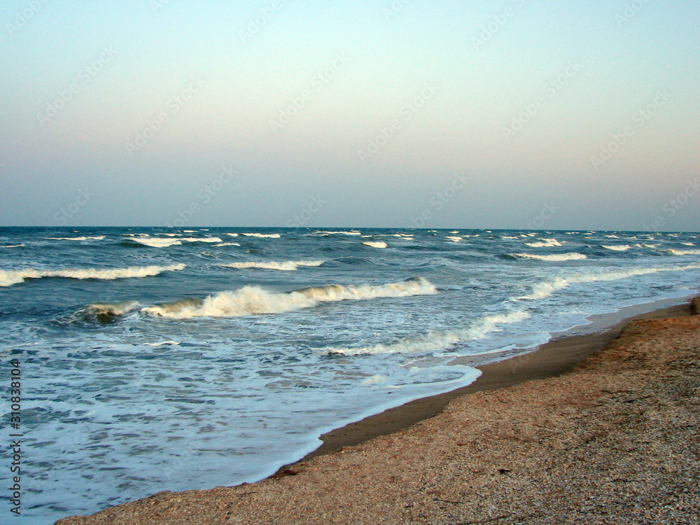 Obraz premium Landscape of snow-white foam of coastal sea waves gently stroking the sandy shore, under the rainbow colors of the sunset.
