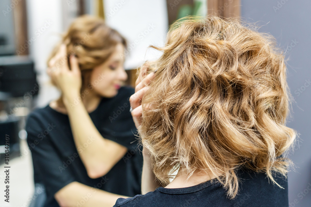 Naklejka premium Beautiful woman is touching her hair, checking her hairstyle in the mirror at beauty salon, copy space.
