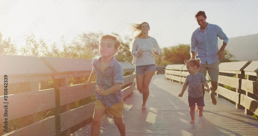 Young happy smiling toddler boys running with their mom and dad on a coastal boardwalk at sunset, happy parents and their children running outdoors together at sunset, happy parenthood moments