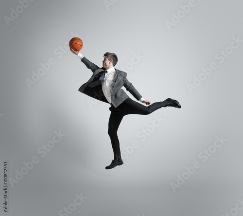 Back in college - it's never too late for being sport star. Man in office clothes traines in basketball on grey background. Unusual look for businessman in motion, action. Sport, healthy lifestyle. photo