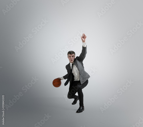 Back in college - it's never too late for being sport star. Man in office clothes traines in basketball on grey background. Unusual look for businessman in motion, action. Sport, healthy lifestyle. photo