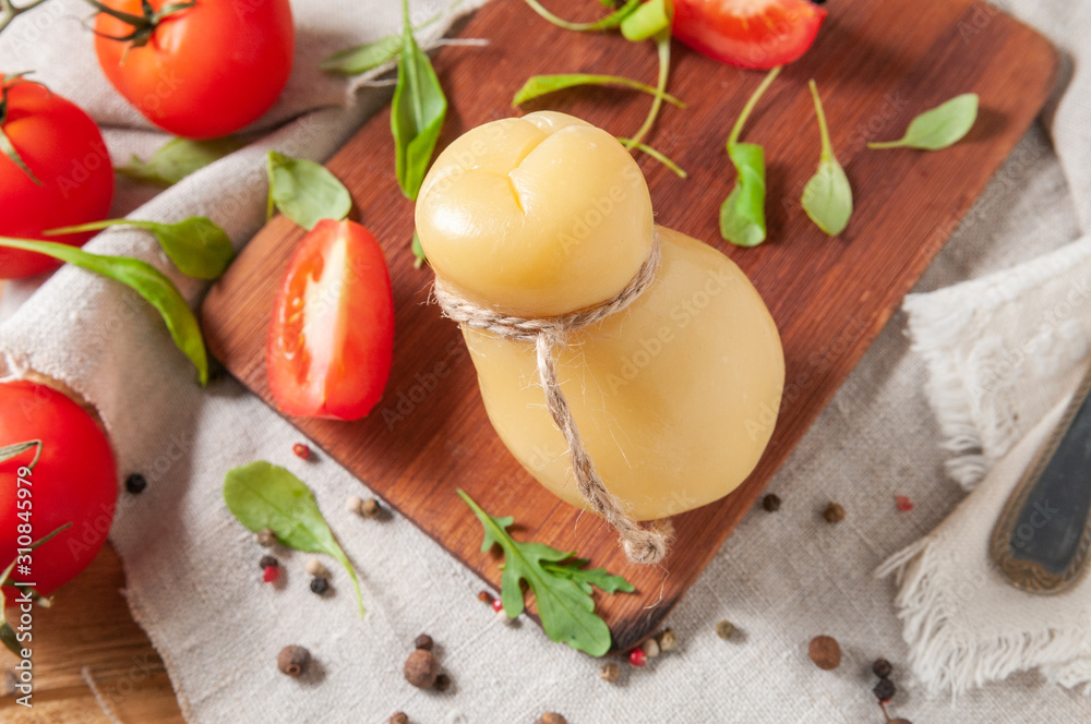 Caciocavallo cheese on a rectangular wooden board. Decorated with arugula, spices and tomatoes. Background gray linen fabric. Close-up. Caciocavallo cheese on a rectangular wooden board. Decorated with arugula, spices and tomatoes. Background gray linen fabric. Close-up.
