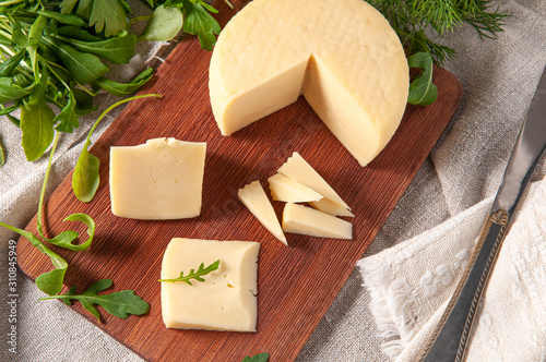 Caciotta cheese is sliced. On a rectangular wooden board. Decorated with arugula, parsley, cilantro. Background gray linen fabric. View from above. photo