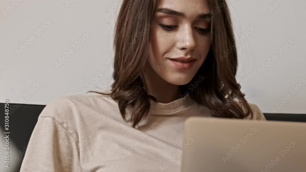 Happy brunette woman using laptop computer while sitting on sofa indoors Happy brunette woman using laptop computer while sitting on sofa indoors