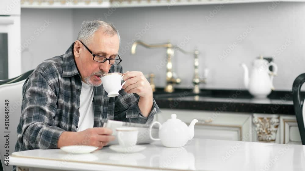 Confident middle aged bearded man drinking tea during reading newspaper at white kitchen interior Confident middle aged bearded man drinking tea during reading newspaper at white kitchen interior