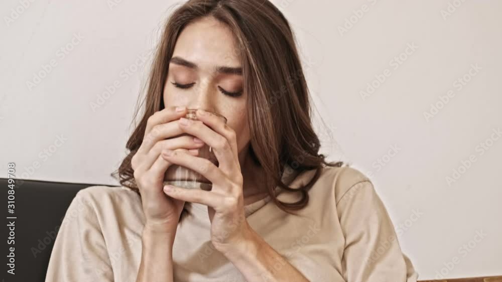 Close up view of pleased pretty brunette woman sniffing cocoa flavor and enjoying this moment with closed eyes while sitting on sofa indoors Close up view of pleased pretty brunette woman sniffing cocoa flavor and enjoying this moment with closed eyes while sitting on sofa indoors