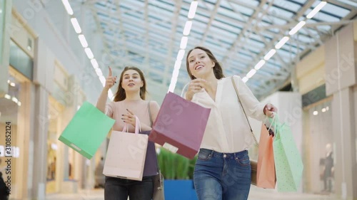 Two excited young women with shopping bags running in slow motion along mall on Black Friday hurrying to make purchase and pointing on stores