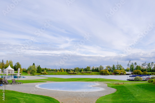 Landscape. View of the beautiful lake and small ornamental ponds covered with paving tiles, lawn with grass, ornamental shrubs and trees, tents and loungers for recreation in the park.