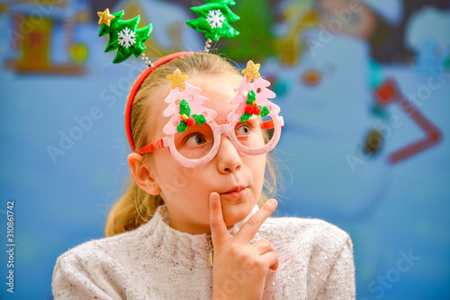 A girl in Christmas clothes, glasses and Christmas trees on her head, looks up thoughtfully in anticipation of a miracle.