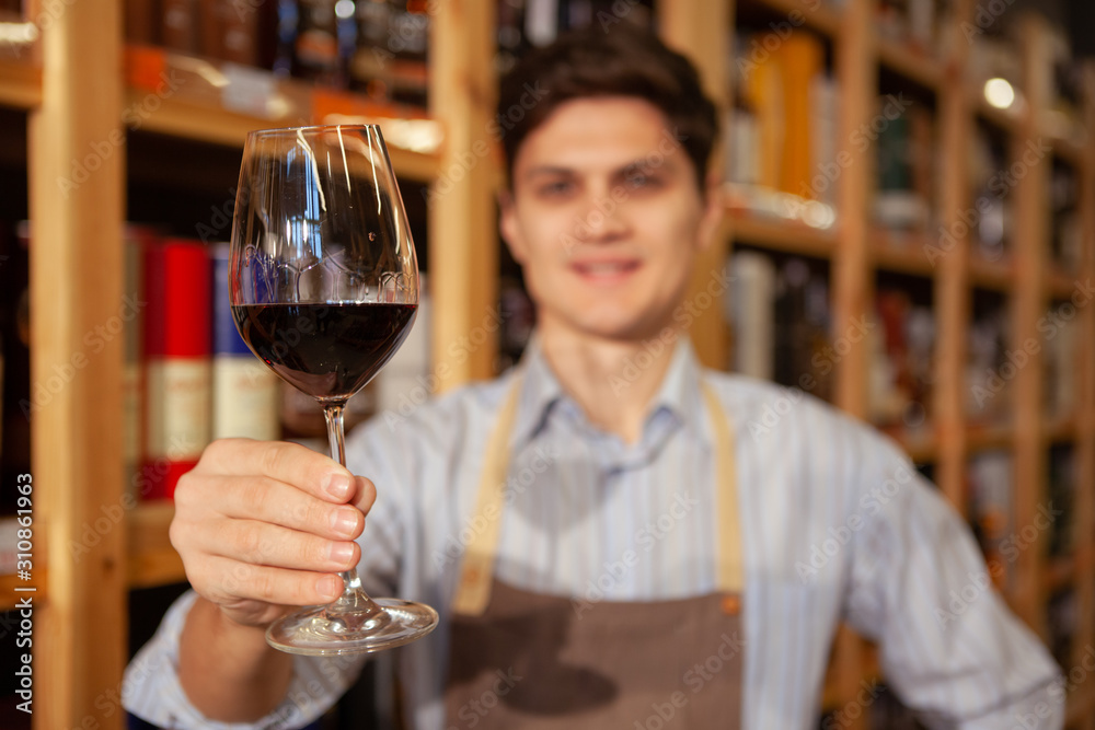 Selective focus on a red wine glass in the hand of cheerful sommelier. Winemaker working at his shop Selective focus on a red wine glass in the hand of cheerful sommelier. Winemaker working at his shop