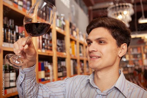 Close up of a cheerful young man tasting wine at wine cellar. Selective focus on red wine glass in the hand of a happy man