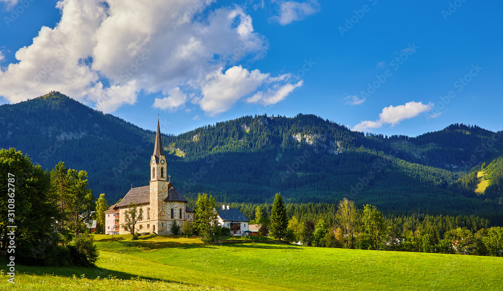 Austria mountain landscape. Traditional church chapel village. Scenic evening sunset panoramic view in austrian Alps mountains. Summer green lawns fields. Knolls covered forest trees. Blue sky clouds.