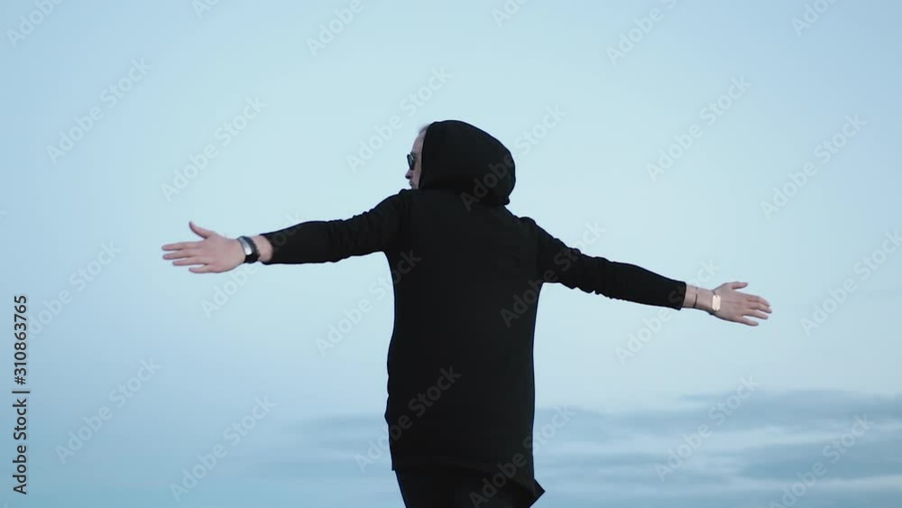 Fashionable young Caucasian guy in a black cardigan with a hood admires nature and fresh air. A man in sunglasses spins with his arms outstretched against a cloudy evening sky. Travel concept Fashionable young Caucasian guy in a black cardigan with a hood admires nature and fresh air. A man in sunglasses spins with his arms outstretched against a cloudy evening sky. Travel concept