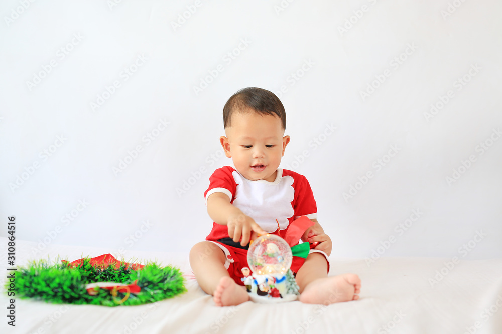 Little Asian baby boy in Santa costume playing snow globe sitting on bed over white background. Merry Christmas and Happy New Year Concept. Little Asian baby boy in Santa costume playing snow globe sitting on bed over white background. Merry Christmas and Happy New Year Concept.