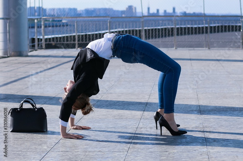slender young woman in black jacket and jeans practice yoga asana bridge pose during break while working day in office