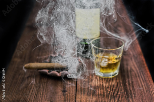  A contrasting and detailed photo of cigar smoke in focus in the foreground. In the background, out of focus on the table, a cigar on the ashtray, a glass of whiskey with ice and a bottle 