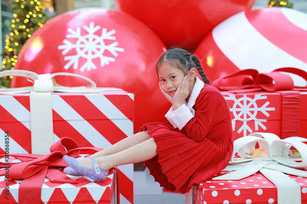 Cute little child girl in red dress poses sitting on big gift in winter season and happy new year festival against christmas background. Stock Adobe Stock