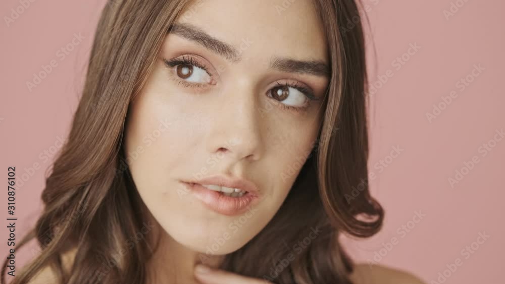 A close-up view of young coquettish woman is biting her lower lip while looking to the camera isolated over pink background